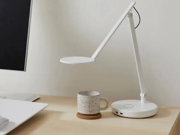 A white adjustable desk lamp with a circular head and base sits on a light wood desk next to a speckled ceramic mug and a computer monitor.